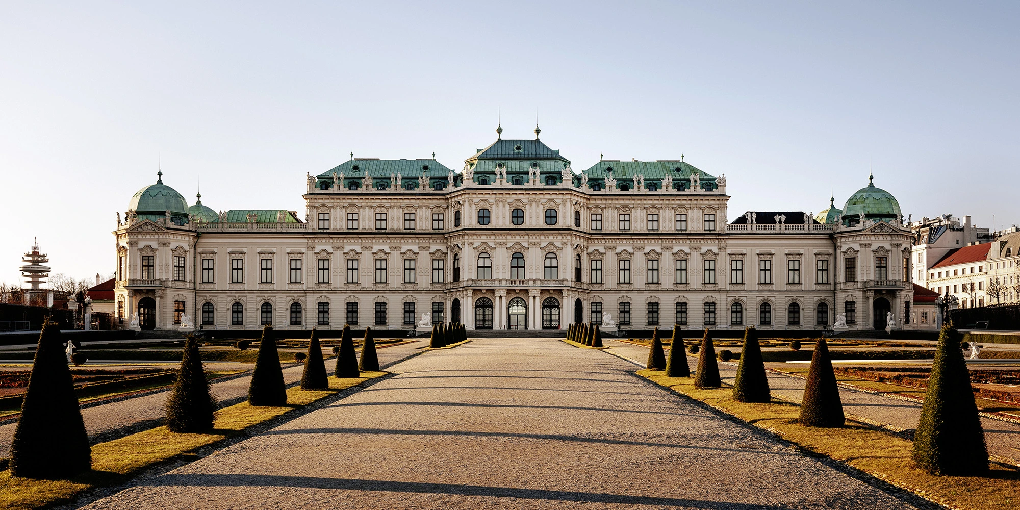 Frontansicht des Oberen Belvedere in Wien bei tiefstehender Sonne, mit symmetrisch angelegtem barocken Garten, kegelförmig beschnittenen Buchsbäumen und klassizistischer Architektur mit grünem Kupferdach und kunstvoll dekorierter Fassade.