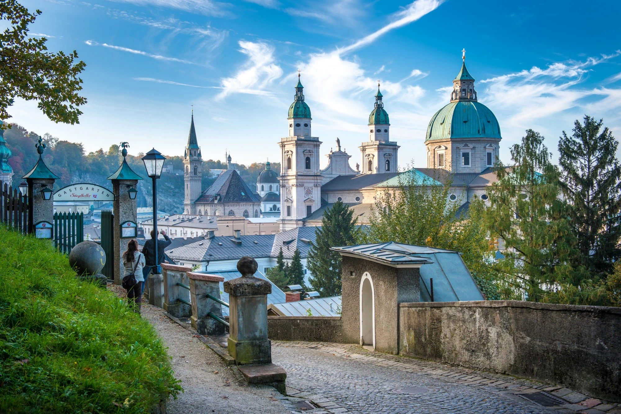 Blick von einem romantischen Fußweg mit Gastgarten-Eingang auf die barocken Türme des Salzburger Doms und umliegende Kirchen vor herbstlicher Hügellandschaft.