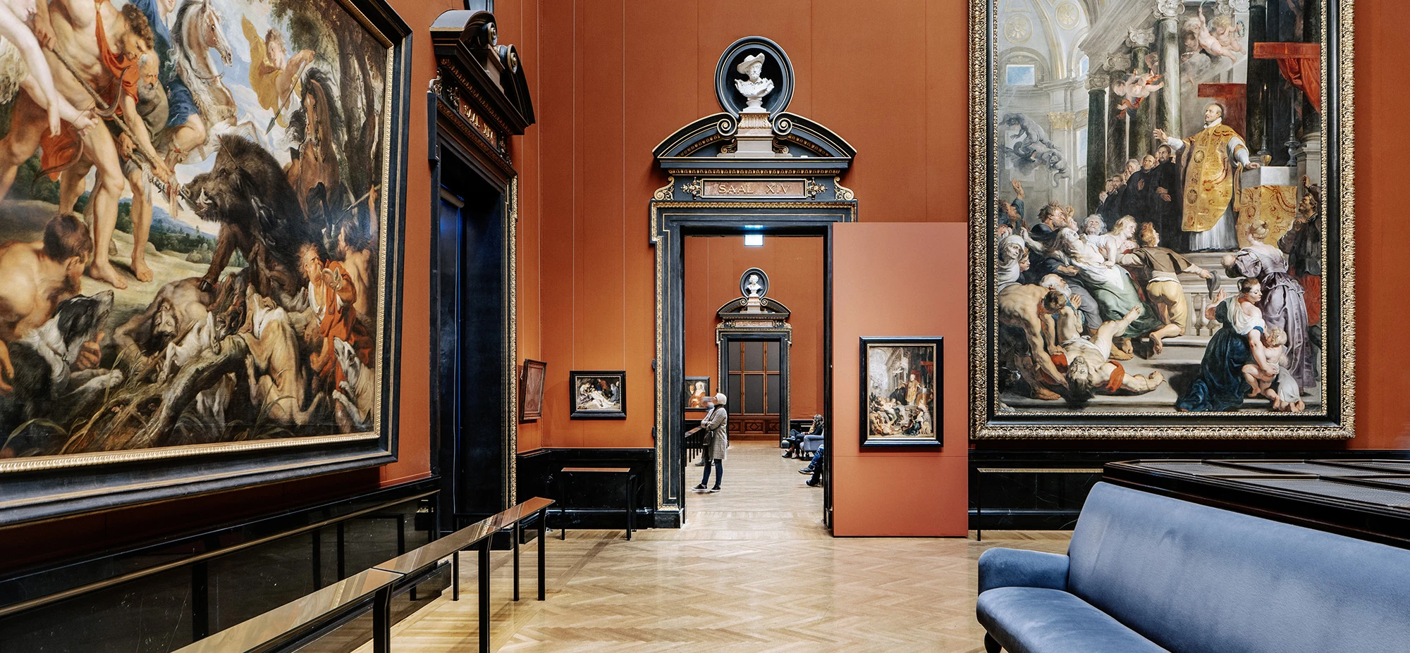 Prunkvoller Museumsraum mit rotem Wandanstrich, großen Barockgemälden in reich verzierten Rahmen, klassizistischen Türeinfassungen mit Büsten und goldenen Ornamenten