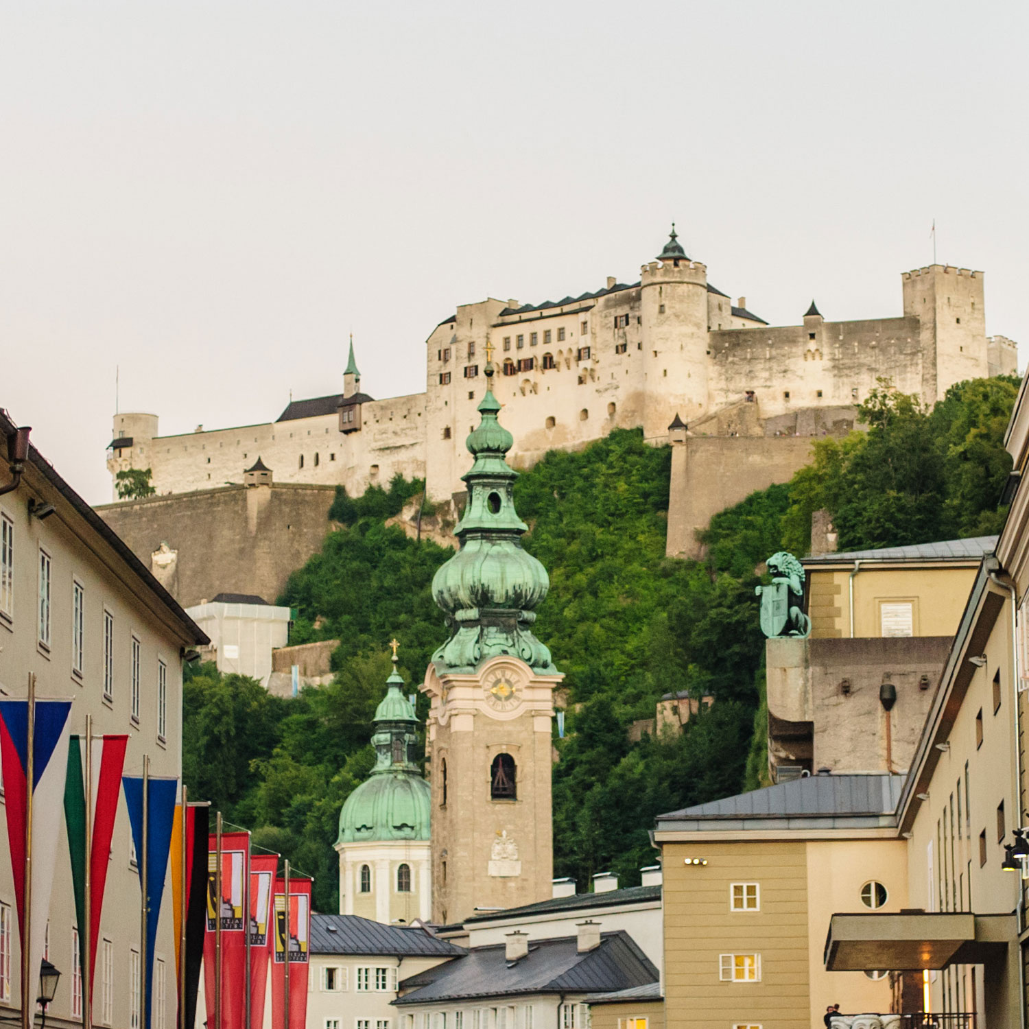 Blick auf die Festung Hohensalzburg, die imposant über den Dächern der Altstadt von Salzburg thront. Im Vordergrund barocke Kirchtürme und bunte Festspiel-Fahnen.