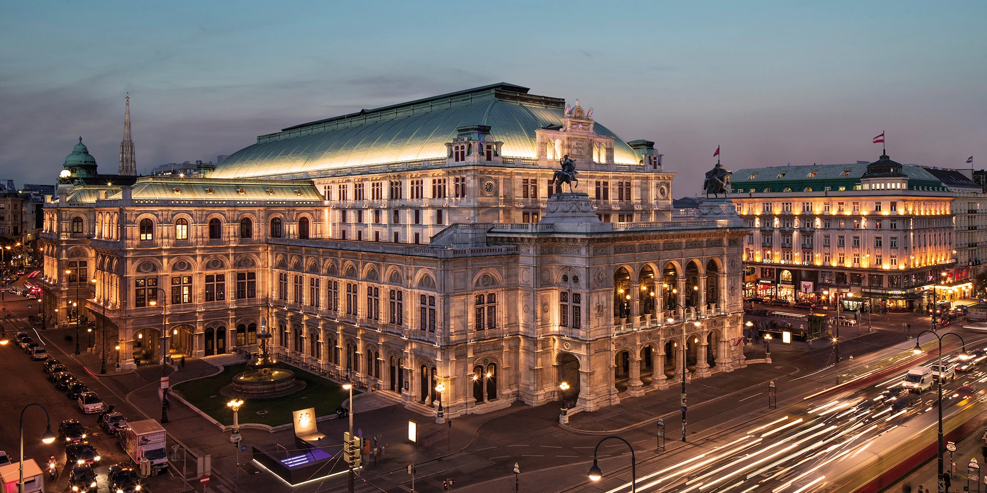 Die Wiener Staatsoper in abendlicher Beleuchtung, majestätisch an der Ringstraße gelegen, mit eindrucksvoller Architektur und regem Verkehr im Vordergrund.