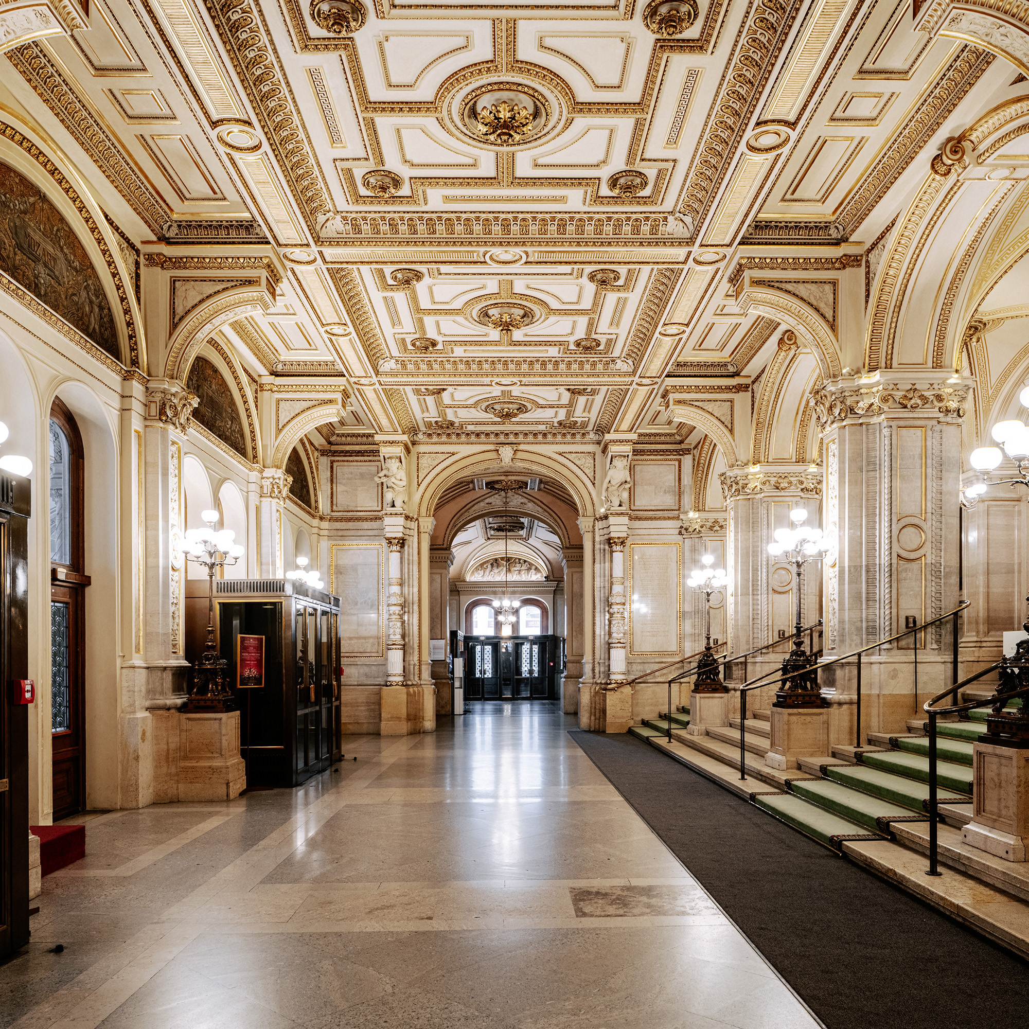 Prächtiges Foyer mit reich verzierter Kassettendecke, klassischen Rundbögen und eleganten Treppenaufgängen, beleuchtet von stilvollen Wand- und Deckenleuchten.