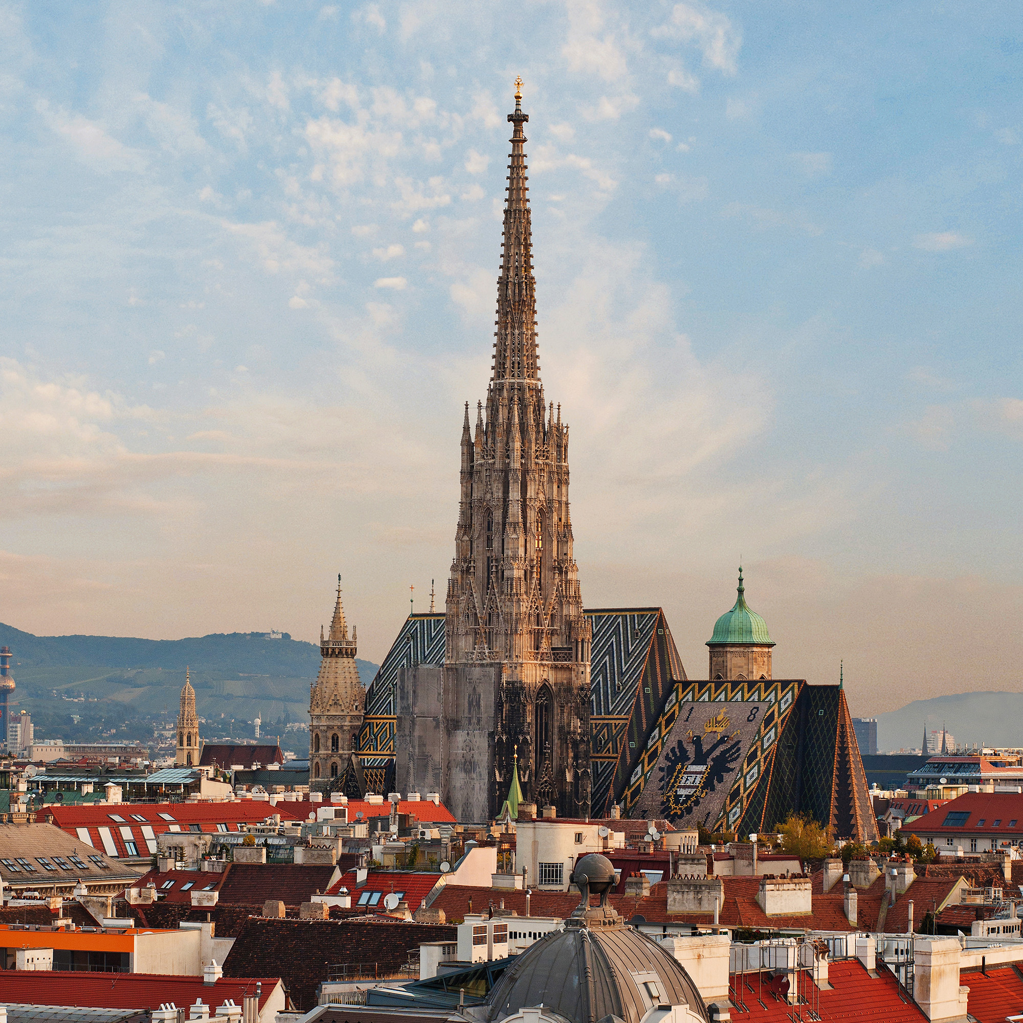 Panorama der Wiener Innenstadt mit dem Stephansdom im Zentrum; die markante gotische Turmspitze ragt über die Stadt, das bunte Ziegeldach mit dem Doppeladler-Motiv ist gut sichtbar, im Hintergrund sanfte Hügel und ein Himmel mit leichten Wolken.