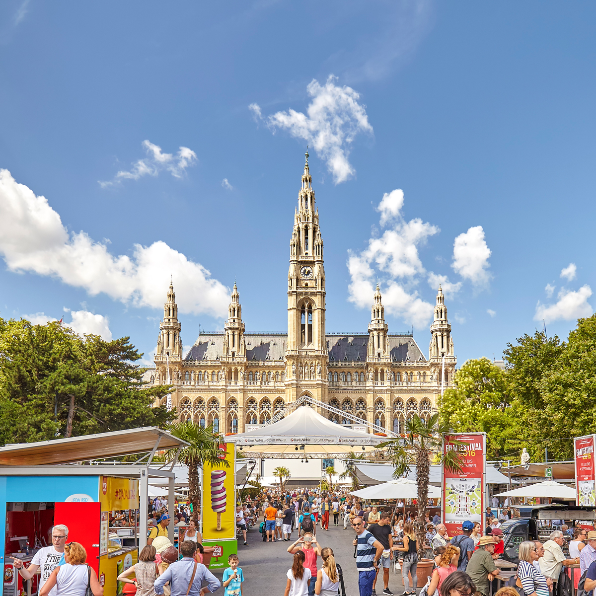 Wiener Rathaus bei Sonnenschein, davor sommerliches Fest mit bunten Ständen, Palmen, Eiswerbung und vielen Besuchern in Freizeitkleidung.