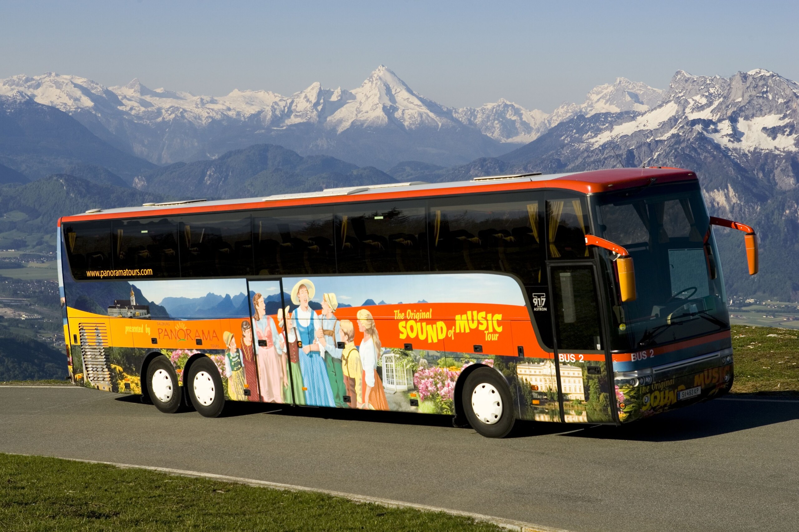 Ein bunter Reisebus der „Original Sound of Music Tour“ vor eindrucksvoller Bergkulisse mit schneebedeckten Gipfeln. Der Bus zeigt Illustrationen aus dem Film „The Sound of Music“ und steht auf einer Straße mit grüner Wiese im Vordergrund.