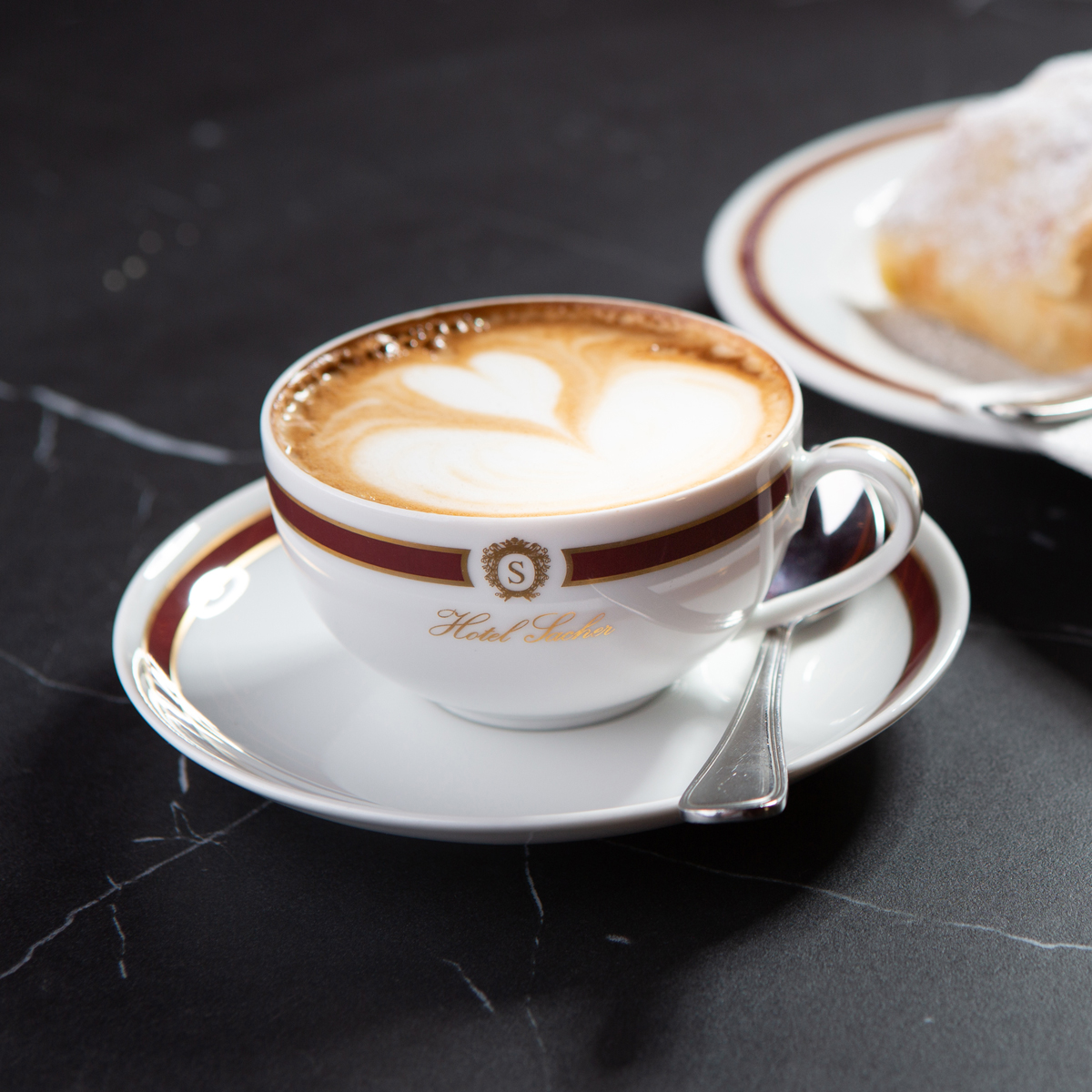 Cappuccino mit Herz-Latte-Art, serviert in einer stilvollen Tasse mit goldverzierter Sacher-Logografie, begleitet von einem Stück klassischem Apfelstrudel auf feinem Porzellan