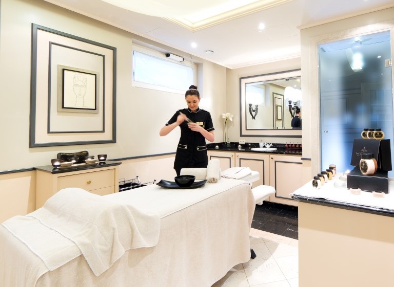 Heller, eleganter Behandlungsraum eines Spas mit Massageliege, auf der eine weiße Decke liegt; eine Mitarbeiterin in schwarzer Uniform bereitet eine Behandlung vor, umgeben von dezentem Dekor und luxuriösen Pflegeprodukten.