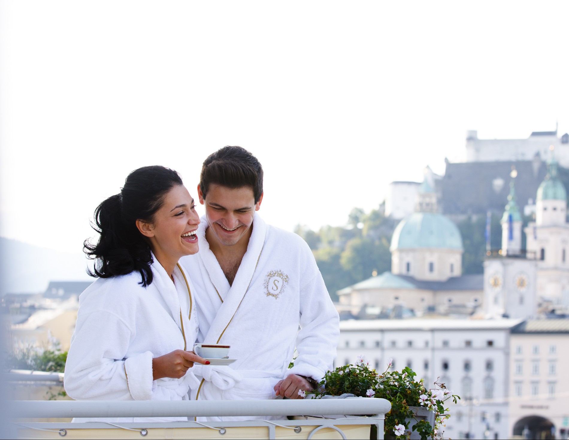 Ein Paar in weißen Bademänteln mit Sacher-Logo auf stehen auf dem Balkon. Beide lachen und genießen gemeinsam eine Tasse Kaffee. Im Hintergrund ist die Silhouette von Salzburg mit der Festung Hohensalzburg und den Kuppeln des Doms zu sehen.