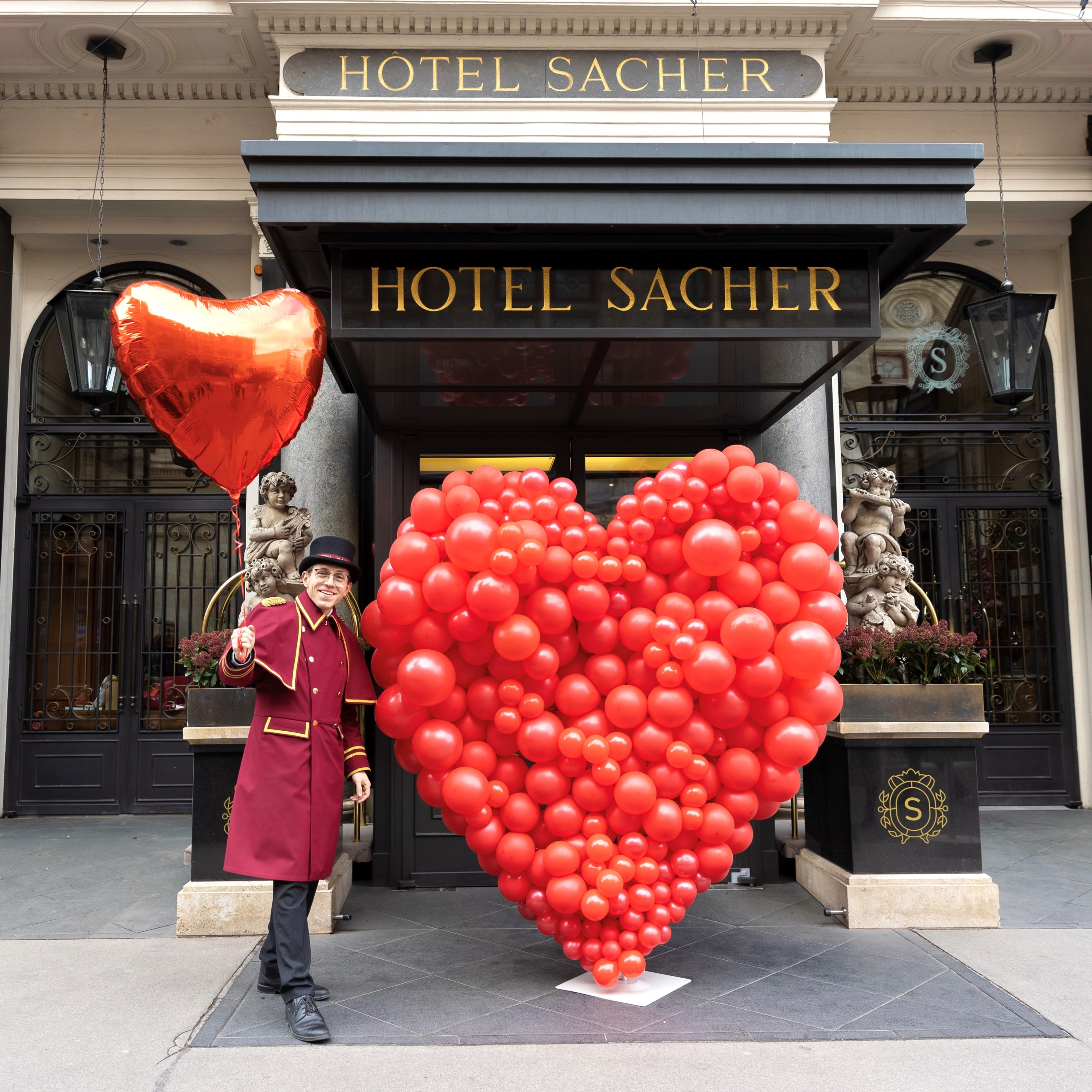 Valentinstag 2023_HSW_002-2 (c) Hotel Sacher Ein Portier in roter Uniform steht vor dem Eingang des Hotel Sacher und hält einen herzförmigen Ballon. Neben ihm ein großes rotes Herz aus vielen kleinen Luftballons. Im Hintergrund die charakteristische Fassade des Hotels.