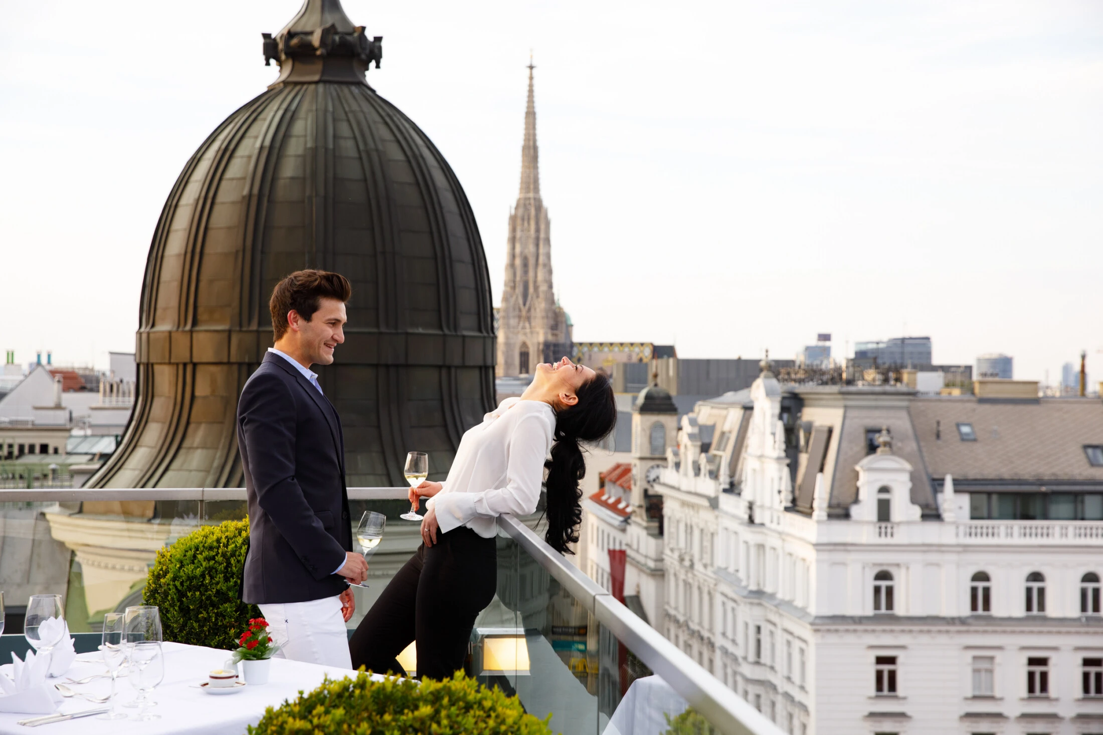 Ein stilvoll gekleidetes Paar genießt ein Glas Wein auf einer eleganten Dachterrasse mit Blick über Wien. Im Hintergrund ragt der imposante Stephansdom empor und schafft gemeinsam mit der historischen Architektur eine romantische Atmosphäre.
