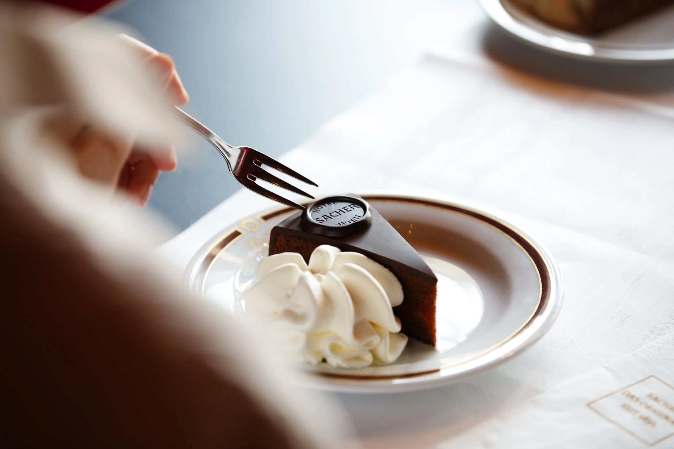 Ein Stück Original Sacher-Torte wird mit einer Gabel angestochen – begleitet von einem Klecks Schlagobers auf einem elegant verzierten Porzellanteller.