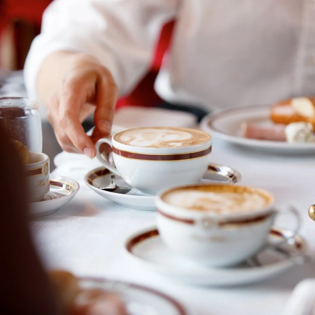 Eine Person greift nach einer eleganten Kaffeetasse mit Latte Art auf einem festlich gedeckten Tisch. Die Szene wirkt einladend und vermittelt gemütlichen Frühstücksgenuss.