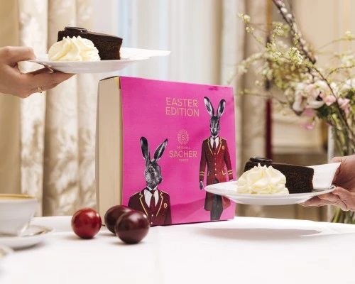 Zwei Hände servieren jeweils ein Stück der Original Sacher-Torte mit Schlagobers auf einem elegant gedeckten Tisch. Im Hintergrund steht die auffällige pinke Osteredition 2022 mit Hasenmotiven – stilvoll umrahmt von Frühlingsblüten und Ostereiern.