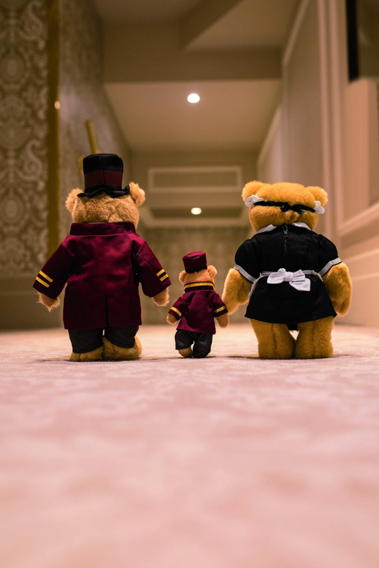 Drei Teddybären im Hotel-Sacher-Look schreiten Hand in Hand einen edlen, weich beleuchteten Hotelgang entlang.