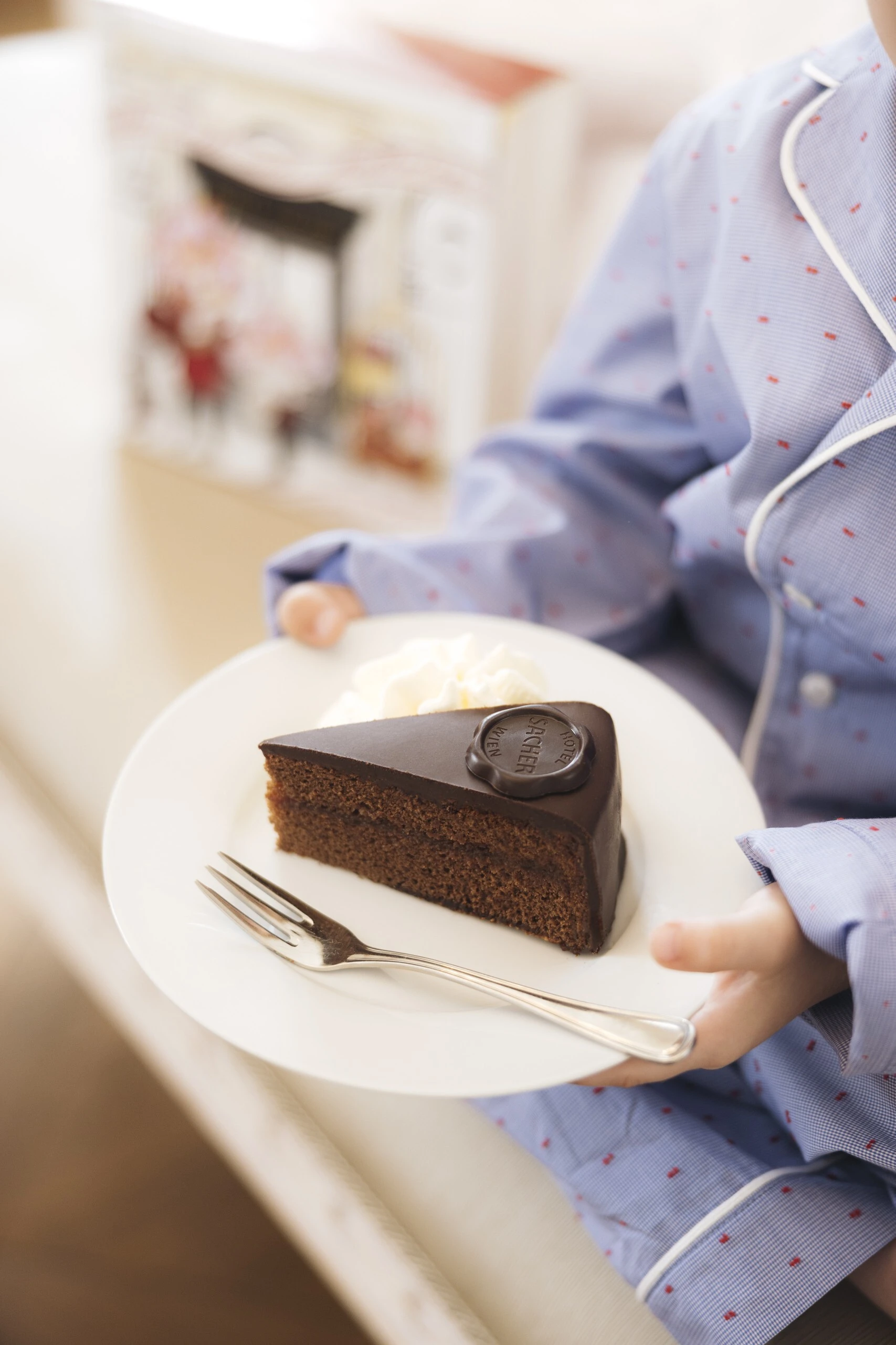 Ein Kind im blauen Pyjama hält einen Teller mit einem Stück Original Sacher-Torte und einem Klecks Schlagobers. Im Hintergrund ist eine festliche Geschenkverpackung vom Hotel Sacher sichtbar