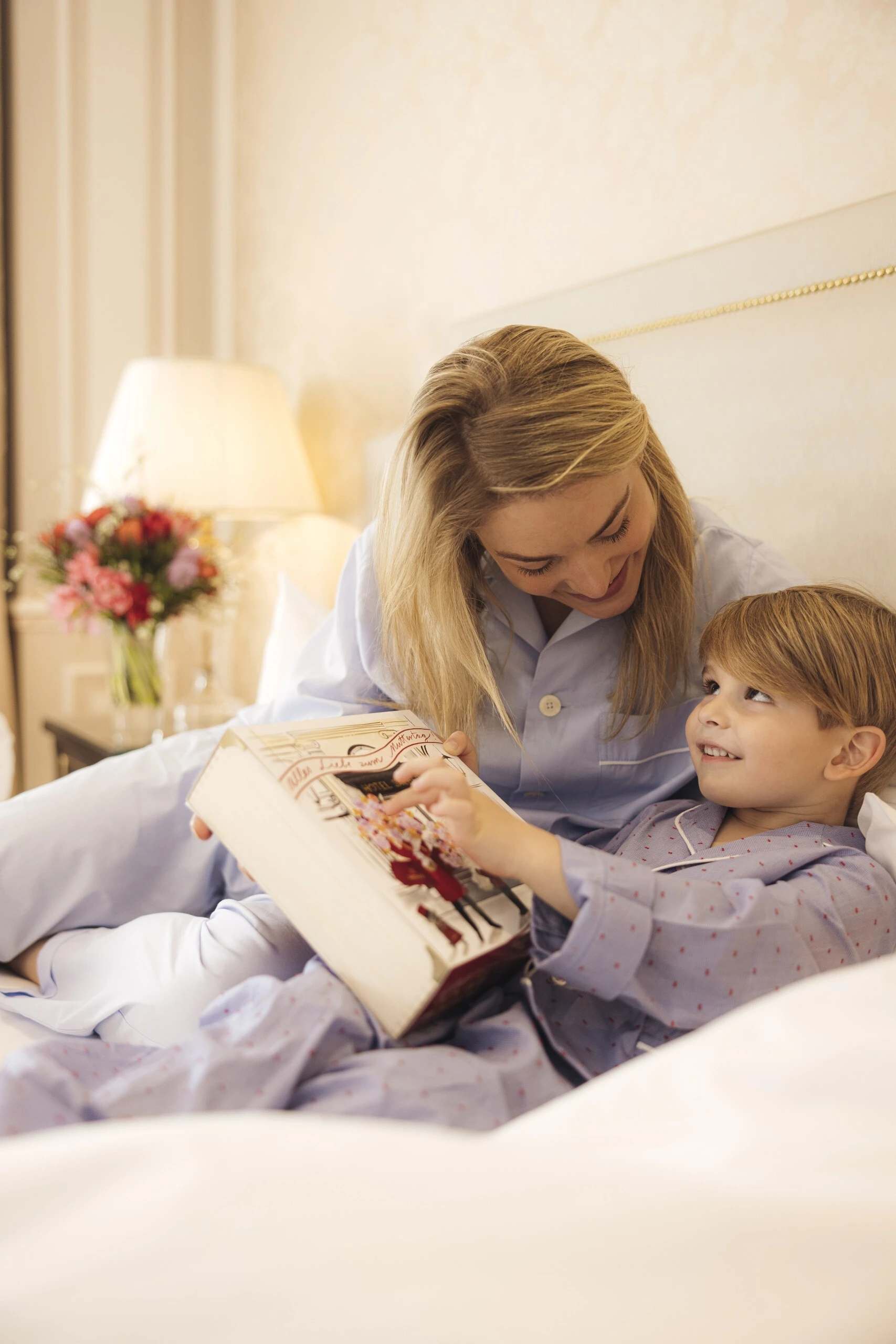 Eine Mutter und ihr Sohn sitzen gemeinsam im Bett, tragen beide Pyjamas und betrachten lächelnd eine dekorative Muttertagsbox von Hotel Sacher. Die Szene wirkt liebevoll und gemütlich.