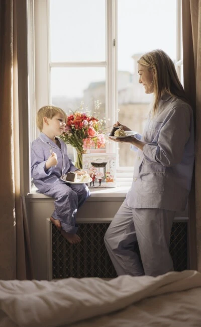 Liebevollen Moment zwischen Mutter und Kind im Hotel Sacher ein. Beide stehen im sanften Morgenlicht am Fenster, genießen gemeinsam ein Stück der legendären Original Sacher-Torte.