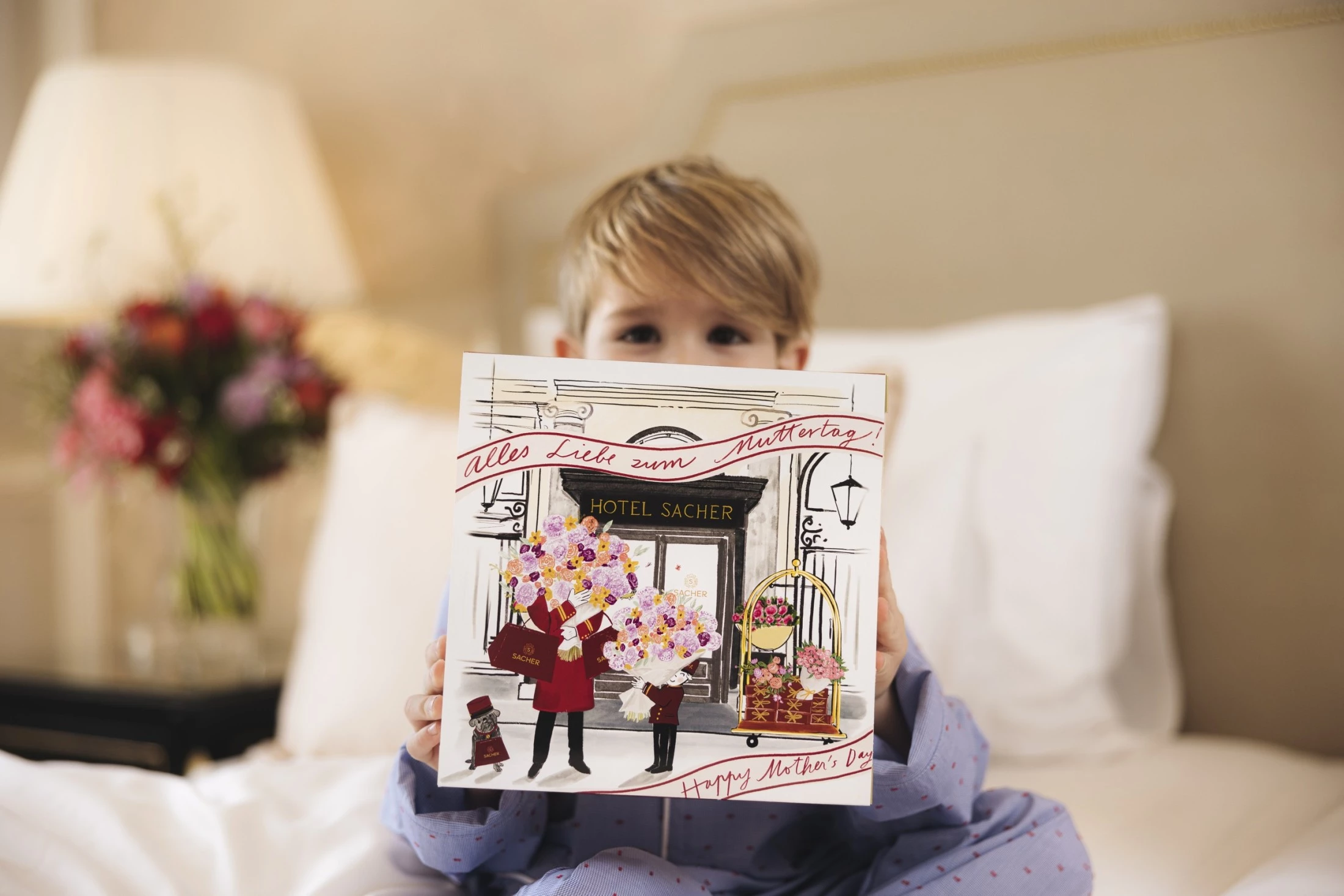 Ein kleiner Junge im Pyjama hält stolz eine zauberhafte Muttertagsbox vom Hotel Sacher in die Kamera – ein Geschenk voller Herzlichkeit und Vorfreude, das mit dem handgezeichneten Motiv auf der Verpackung die besondere Wertschätzung für Mütter stilvoll unterstreicht.