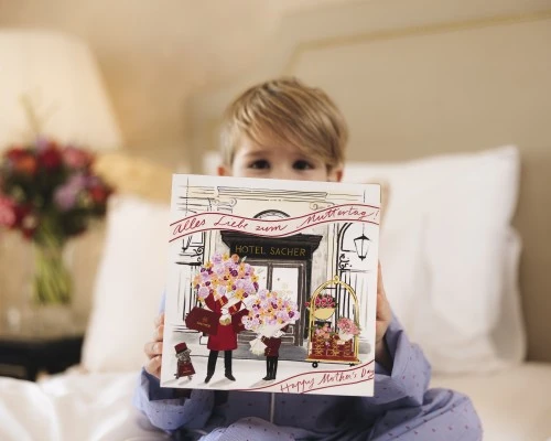 Ein kleiner Junge im Pyjama hält stolz eine zauberhafte Muttertagsbox vom Hotel Sacher in die Kamera – ein Geschenk voller Herzlichkeit und Vorfreude, das mit dem handgezeichneten Motiv auf der Verpackung die besondere Wertschätzung für Mütter stilvoll unterstreicht.