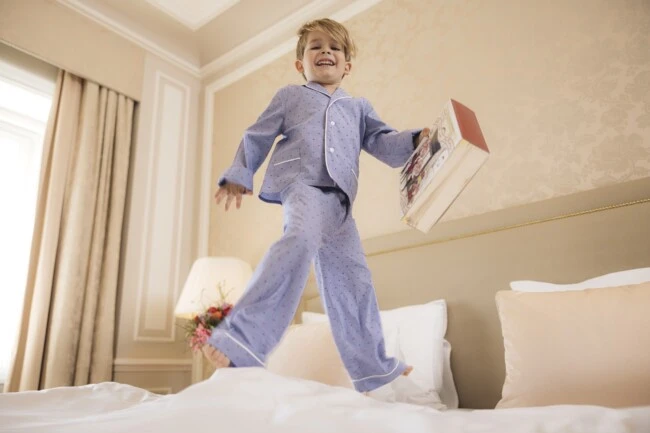 Der Junge springt vergnügt im Hotelbett, trägt hellblaue Pyjamas mit kleinen roten Punkten und hält stolz eine liebevoll gestaltete Muttertags-Sacher-Box in der Hand – pure Vorfreude auf ein süßes Geschenk.