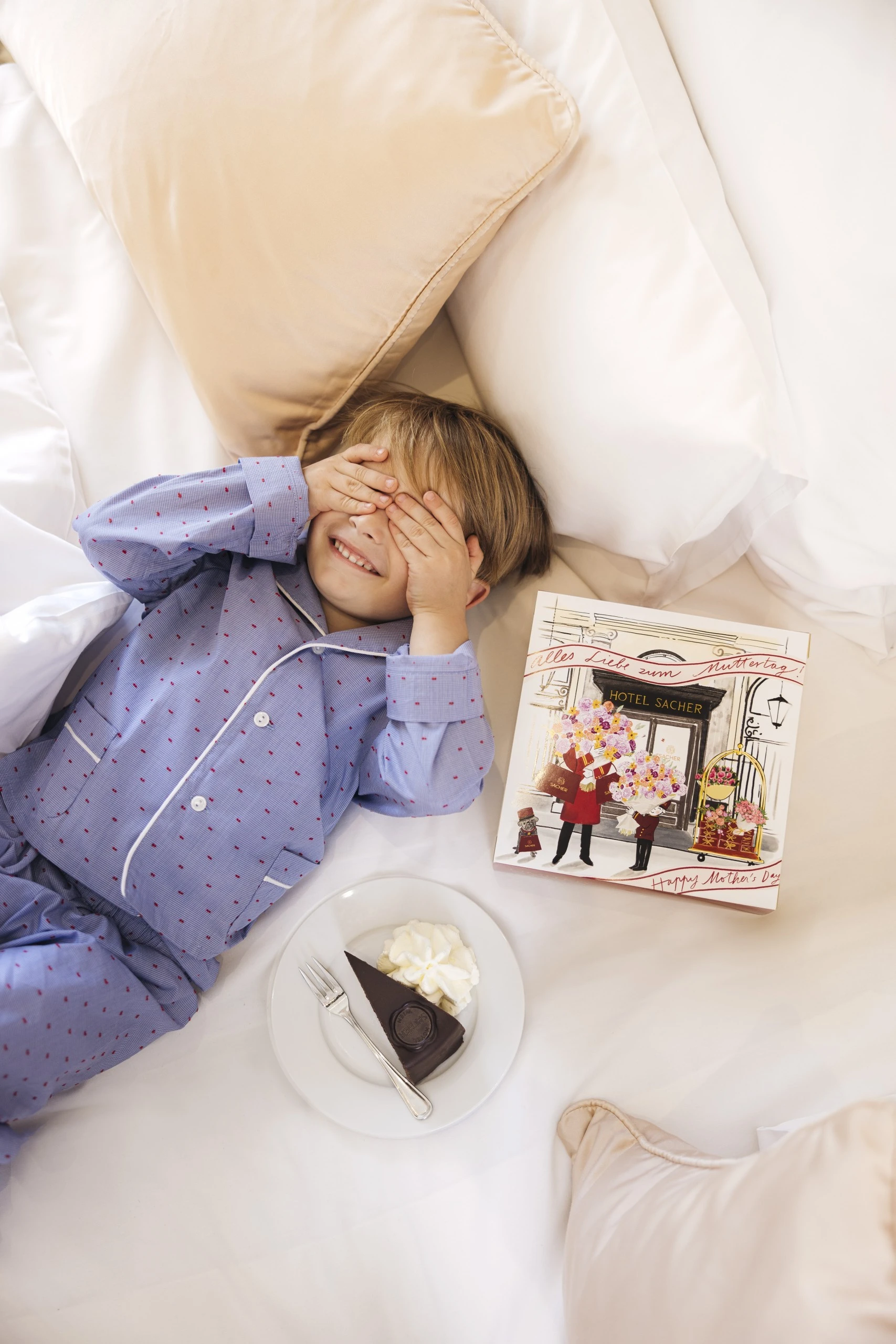 Ein Junge in blauen Pyjamas liegt lachend im Bett, die Augen mit den Händen bedeckt; neben ihm eine Muttertags-Geschenkbox vom Hotel Sacher und ein Teller mit Original Sacher-Torte und Schlagobers.