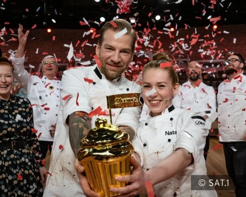 Zwei Köche in weißer Uniform, umgeben von Teammitgliedern und rotem Konfetti, halten strahlend einen goldenen Pokal in Form eines Cupcakes; im Hintergrund festliche Stimmung auf einer Kochshow-Bühne.
