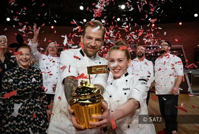 Hotel Sacher Wien, das Große Backen Zwei Köche in weißer Uniform, umgeben von Teammitgliedern und rotem Konfetti, halten strahlend einen goldenen Pokal in Form eines Cupcakes; im Hintergrund festliche Stimmung auf einer Kochshow-Bühne.