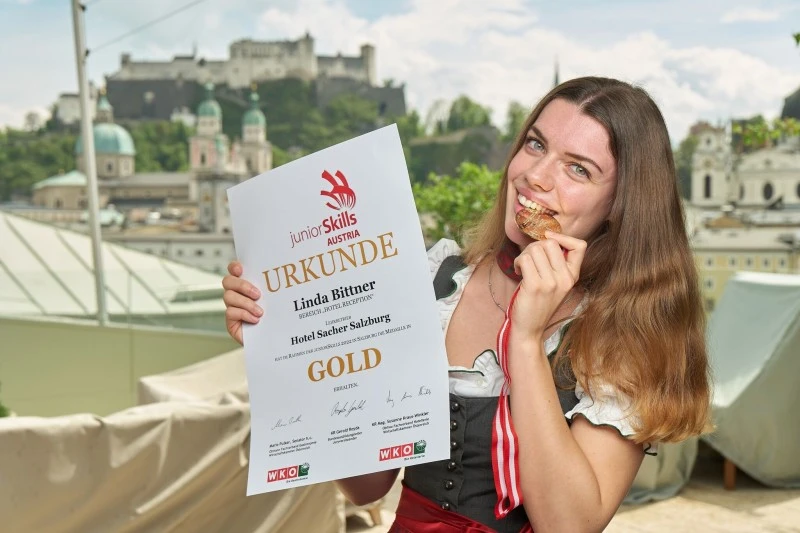 Hotel Sacher Salzburg, Junior Skills Junge Frau in Tracht hält stolz eine Gold-Urkunde der Junior Skills Austria für den Bereich „Hotel Reception“, Ausbildungsbetrieb Hotel Sacher Salzburg, und beißt lachend auf ihre Goldmedaille; im Hintergrund die Altstadt von Salzburg mit Festung und Kirchen.