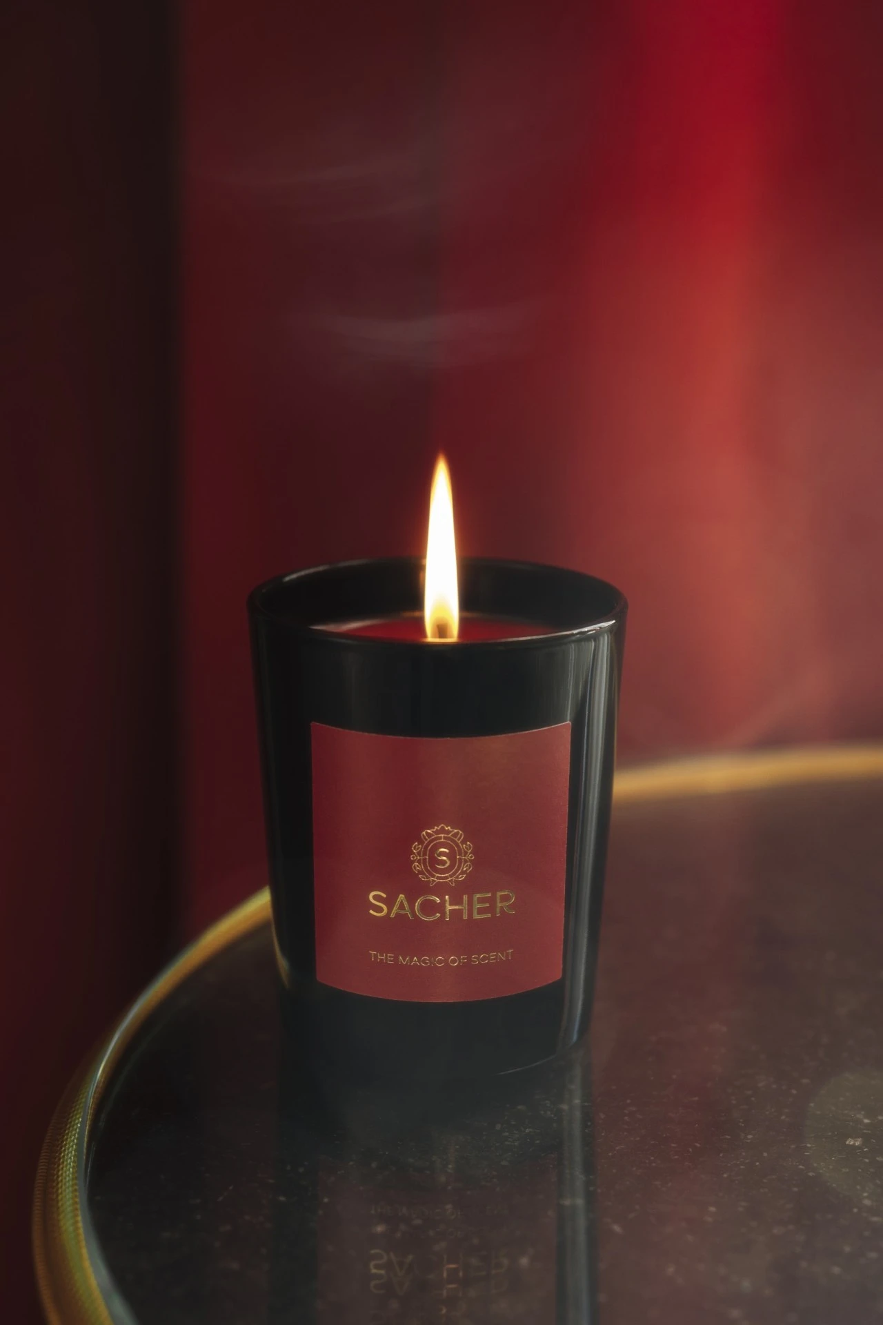 Brennende Sacher-Duftkerze in schwarzem Glas mit rotem Etikett auf einem runden, dunklen Tisch; im Hintergrund sanftes, warmes Licht und ein intensiver roter Farbverlauf.
