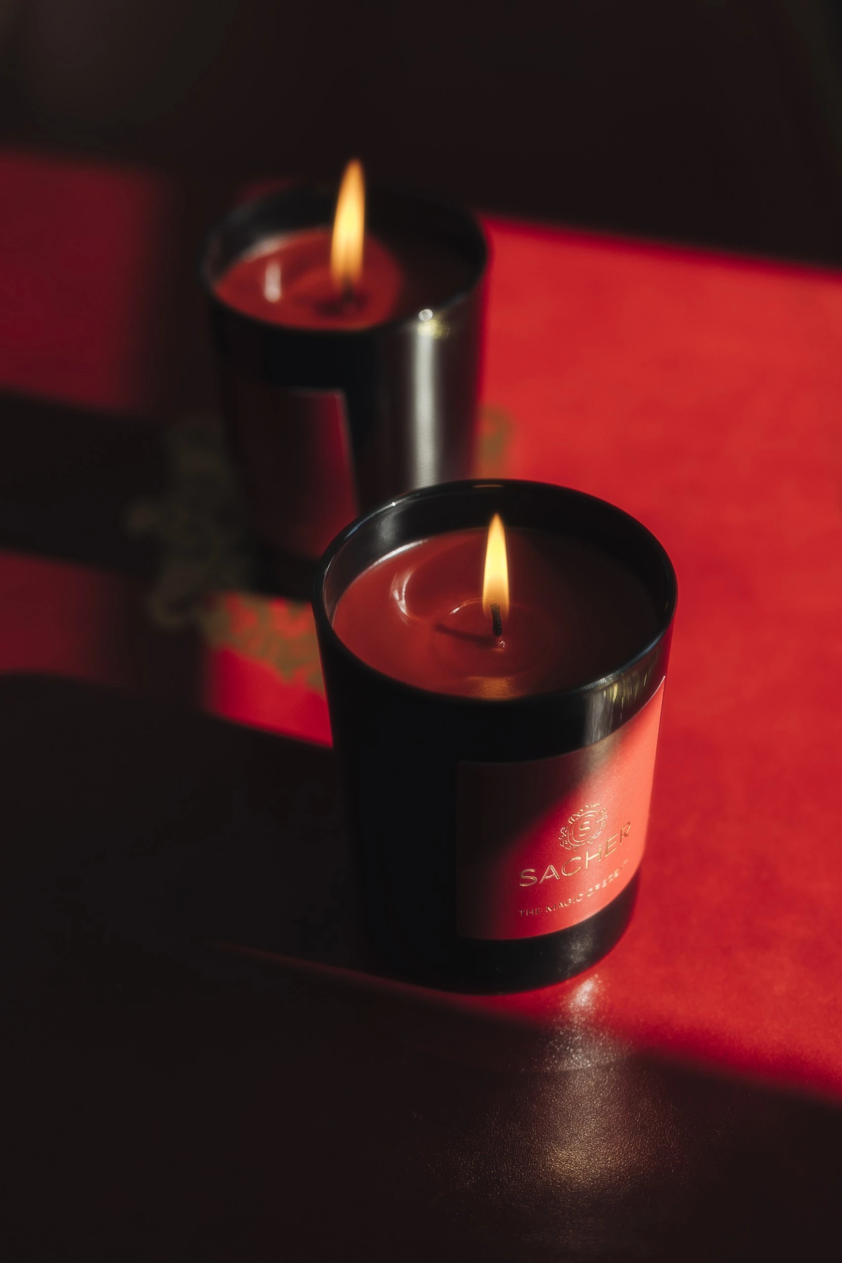 Zwei brennende Sacher-Duftkerzen auf leuchtend rotem Untergrund, warme Lichtstrahlen betonen das glänzende schwarze Glas und die goldene Schrift auf dem roten Etikett.