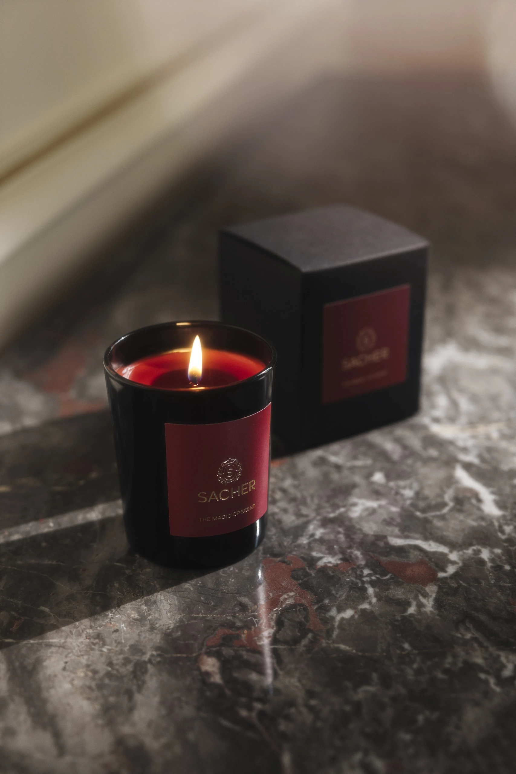 Brennende Duftkerze in schwarzem Glas mit rotem Label und goldener Sacher-Schrift „THE MAGIC OF SCENT“, dahinter die passende schwarze Verpackung; auf edlem Marmor in warmem Licht präsentiert.