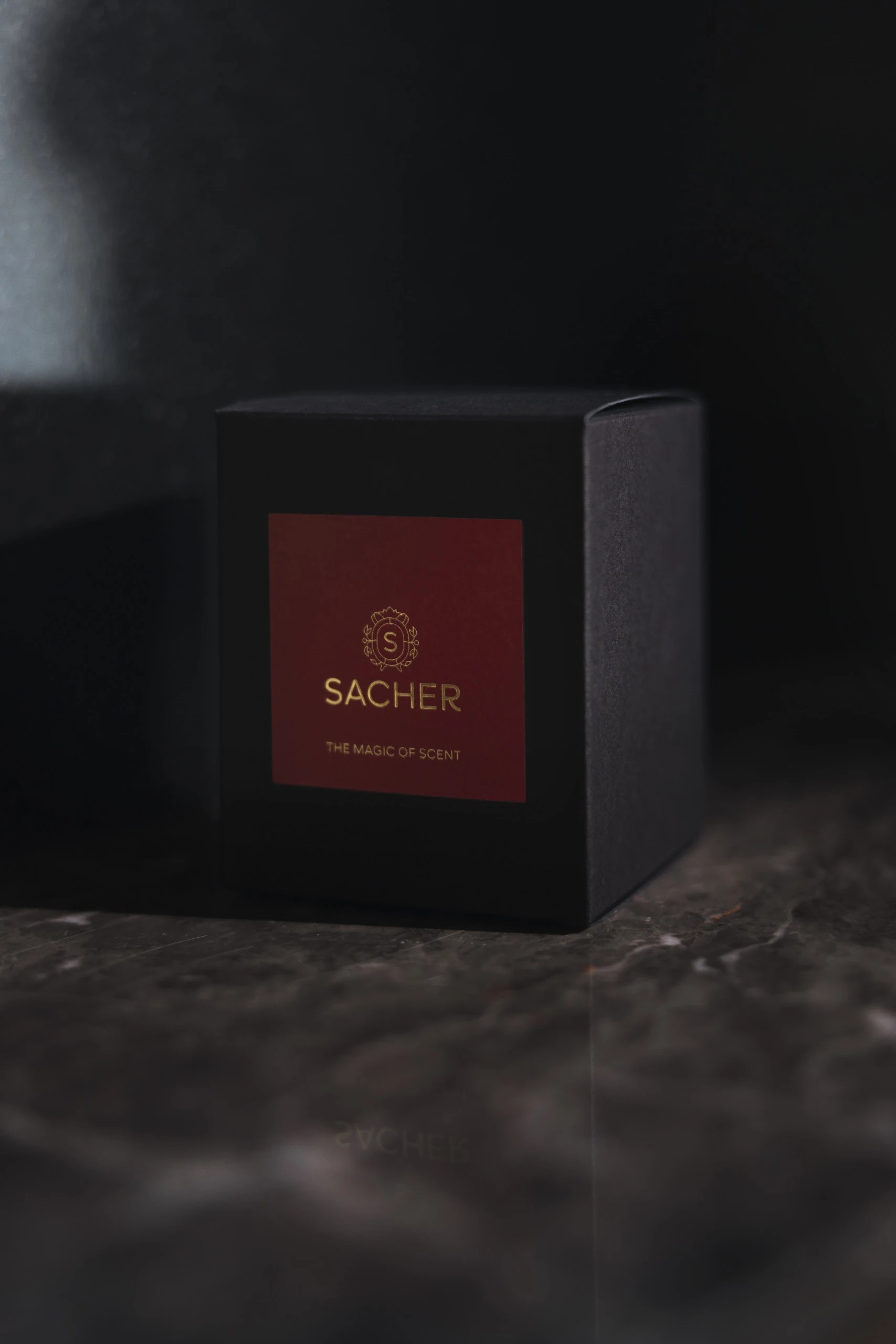 Schwarze Verpackung mit rotem Etikett und goldener Sacher-Aufschrift „THE MAGIC OF SCENT“, elegant in Szene gesetzt auf dunklem Marmor mit weichem Schatten- und Lichtspiel.