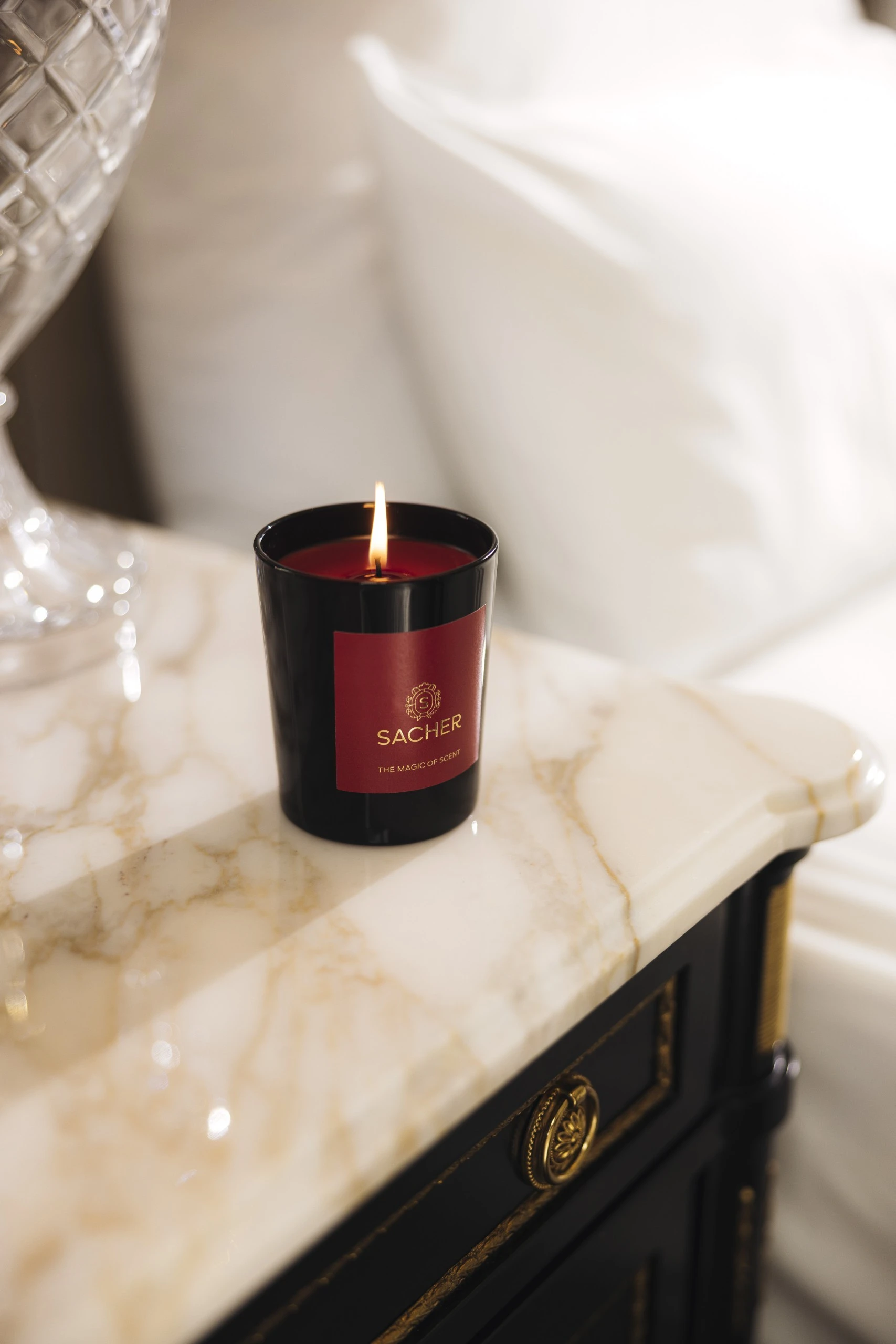 Brennende Duftkerze in schwarzem Glas mit rotem Etikett und goldener Sacher-Aufschrift „The Magic of Scent“, auf hellem Marmor mit funkelndem Hintergrundlicht.
