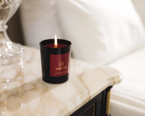 Brennende Duftkerze in schwarzem Glas mit rotem Etikett und goldener Sacher-Aufschrift „The Magic of Scent“, auf hellem Marmor mit funkelndem Hintergrundlicht.