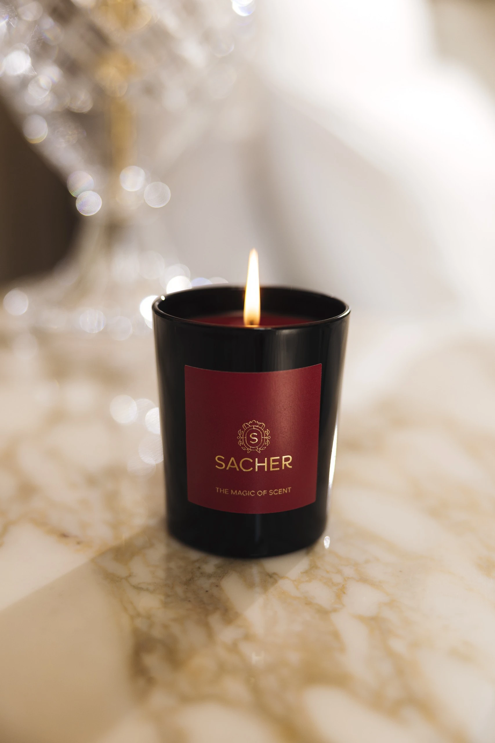 Brennende Duftkerze in schwarzem Glas mit rotem Etikett und goldener Sacher-Aufschrift „The Magic of Scent“, auf hellem Marmor mit funkelndem Hintergrundlicht.