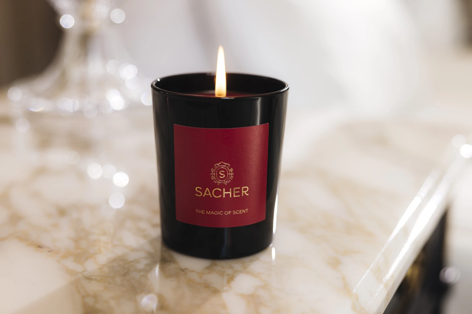 Brennende Duftkerze in schwarzem Glas mit rotem Etikett und goldener Sacher-Aufschrift „The Magic of Scent“, auf hellem Marmor mit funkelndem Hintergrundlicht.