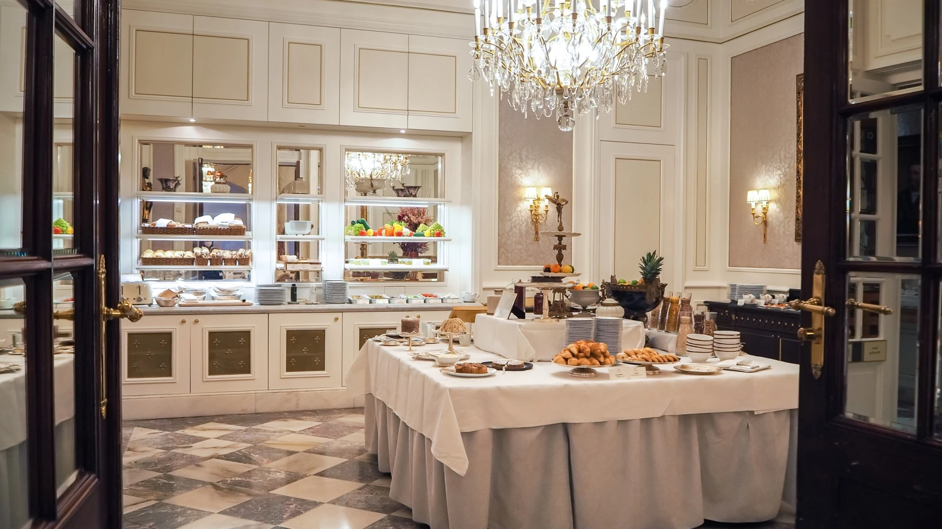 Exklusiver Frühstücksraum mit zentralem Buffet aus frischem Gebäck, Obst und Aufstrichen, umgeben von Marmorboden, hohen Wandvertäfelungen, vergoldeten Details und einem imposanten Kristalllüster.