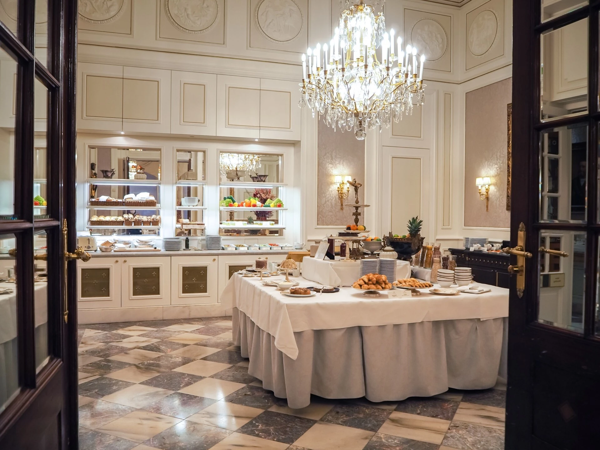 Exklusiver Frühstücksraum mit zentralem Buffet aus frischem Gebäck, Obst und Aufstrichen, umgeben von Marmorboden, hohen Wandvertäfelungen, vergoldeten Details und einem imposanten Kristalllüster.
