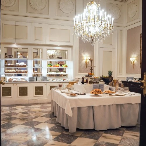 Exklusiver Frühstücksraum mit zentralem Buffet aus frischem Gebäck, Obst und Aufstrichen, umgeben von Marmorboden, hohen Wandvertäfelungen, vergoldeten Details und einem imposanten Kristalllüster.