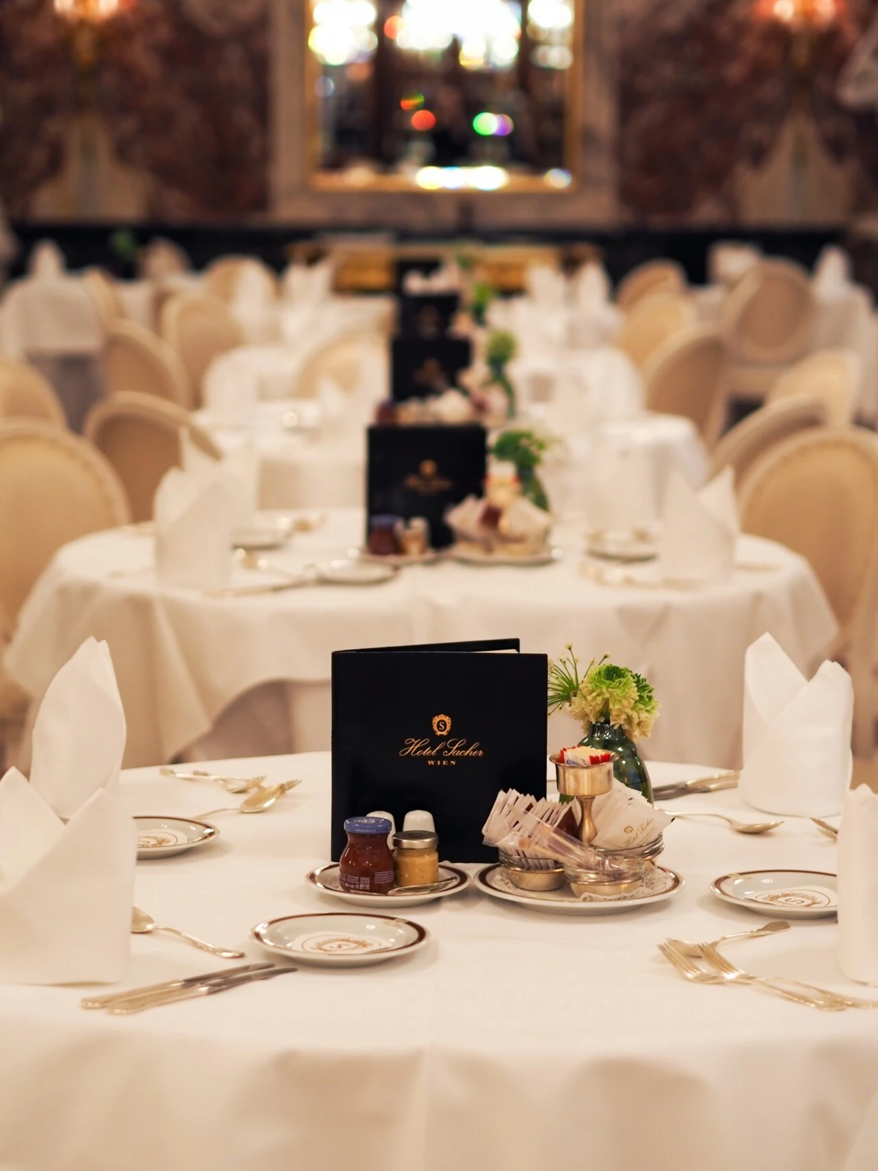 Raffiniert gedeckter Frühstückstisch im Hotel Sacher Wien mit weißer Tischdecke, eleganten Tellern, Besteck, Servietten und schwarzer Speisekarte; im Hintergrund zahlreiche gleichartig vorbereitete Tische in stilvollem Ambiente.