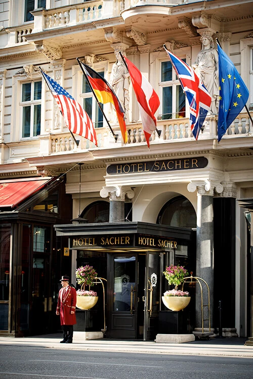 Eingang des Hotel Sacher mit wehenden Nationalflaggen, klassischer Architektur und Portier in roter Uniform.