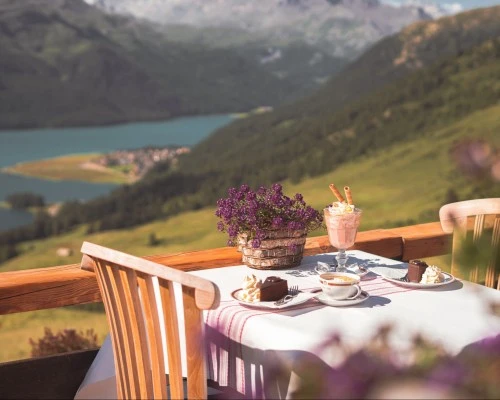 Frühstückstisch auf einer Panorama-Terrasse mit zwei Portionen Original Sacher-Torte, Cappuccino, Eiskaffee mit Waffelröllchen und Blumentopf; im Hintergrund ein malerischer Blick auf Berge, Wälder und einen tiefblauen See.