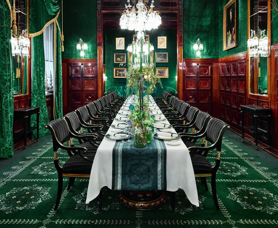 Hotel Sacher Wien, Grüne Bar Prunkvoller Speisesaal mit langem, elegant gedecktem Tisch für 20 Personen, umgeben von schwarzen Polsterstühlen mit Goldakzenten, grün-goldener Teppich und Vorhängen, dunklem Holzvertäfelungen und opulenten Kronleuchtern.