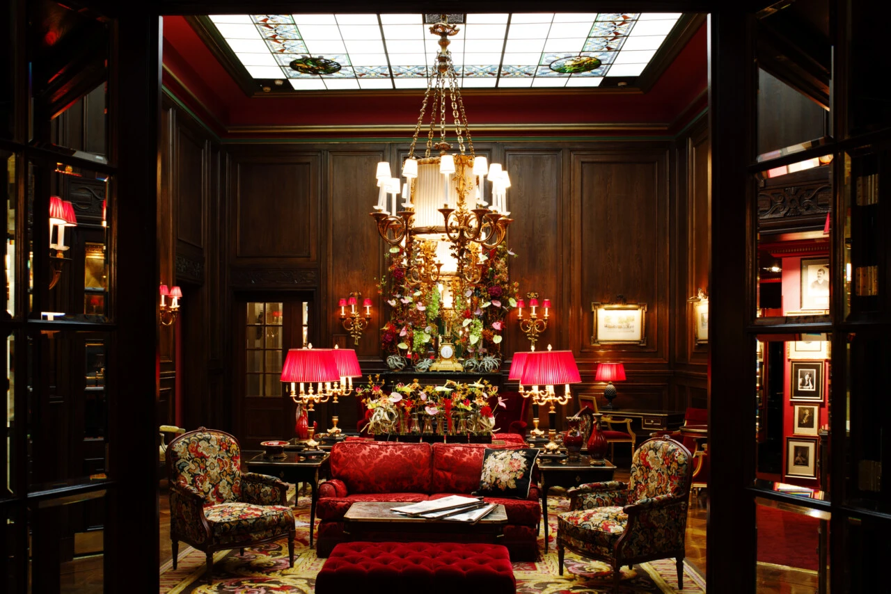 Prunkvoller Salon mit dunkler Holzvertäfelung, üppiger Blumendekoration, opulenten Kronleuchtern und eleganten Möbeln in Rot- und Goldtönen.