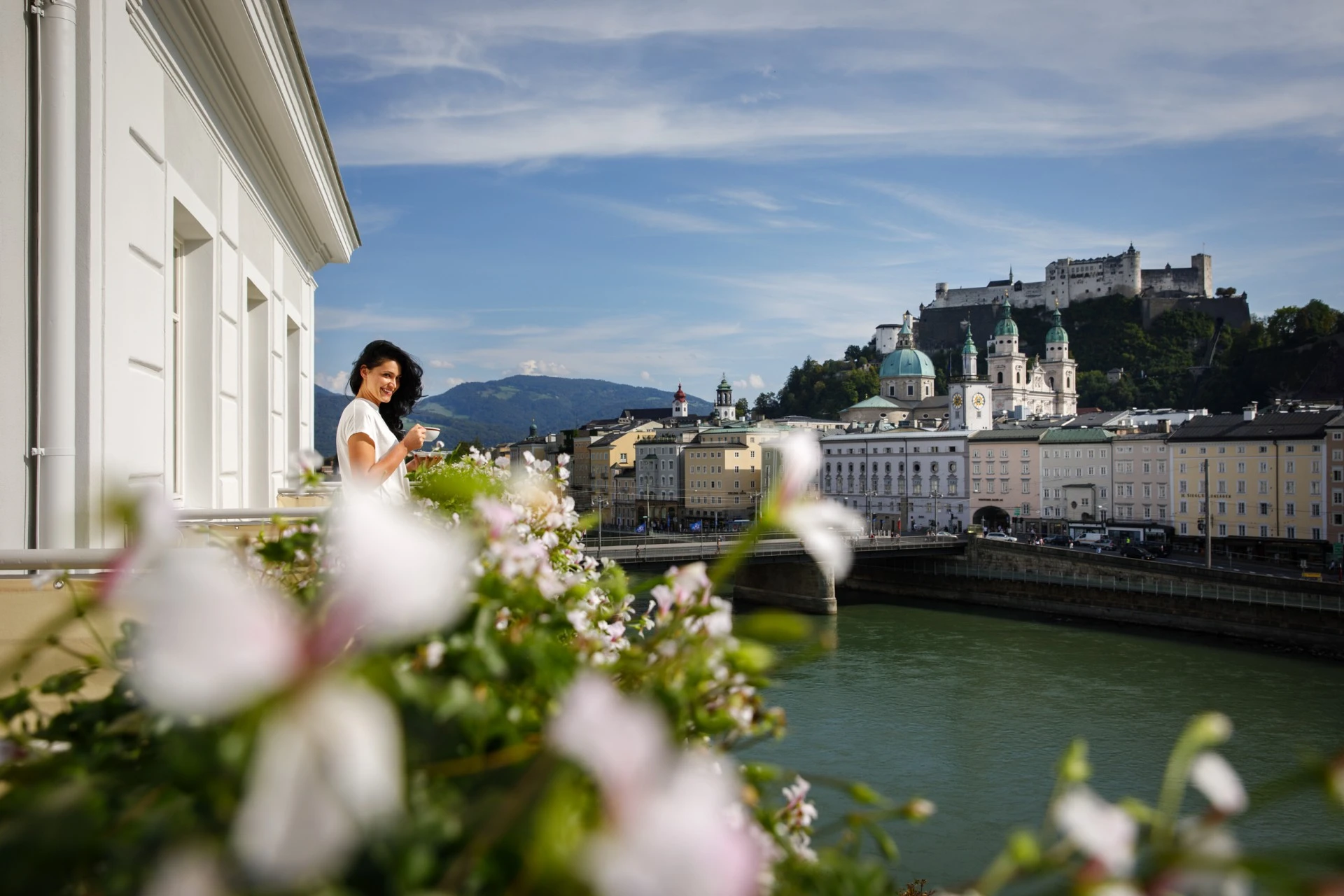 Frau auf einer Terrasse mit Blick auf die Salzburger Altstadt und die Festung Hohensalzburg, umgeben von blühenden Blumen und bei strahlendem Sonnenschein.