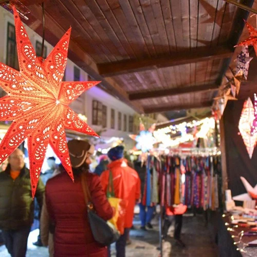 Ein bunter Weihnachtsmarkt mit leuchtenden, dekorativen Sternenlampen, bunten Ständen und Besuchern in Winterkleidung.