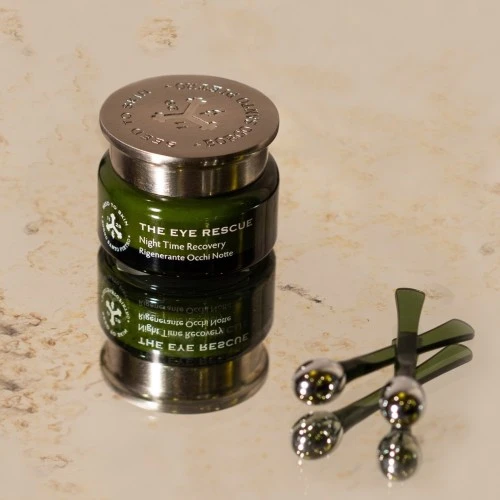 Ein elegantes Glasdöschen mit der Aufschrift „The Eye Rescue – Night Time Recovery“ steht auf einer glänzenden Oberfläche, daneben liegen drei kleine grüne Massagetools.