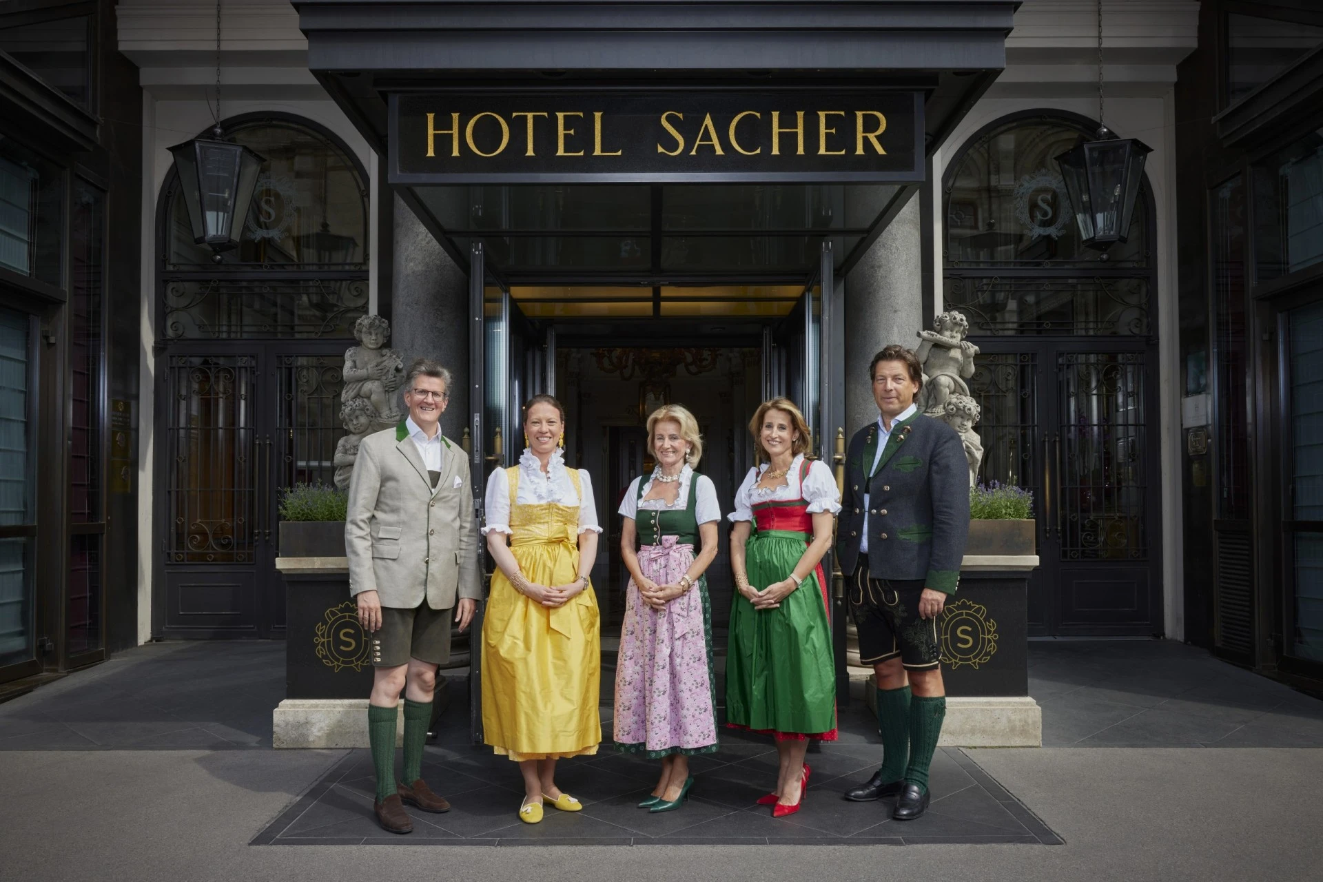 Mitarbeitende in Tracht und Uniformen stehen freundlich vor dem traditionsreichen Hotel Sacher Wien.