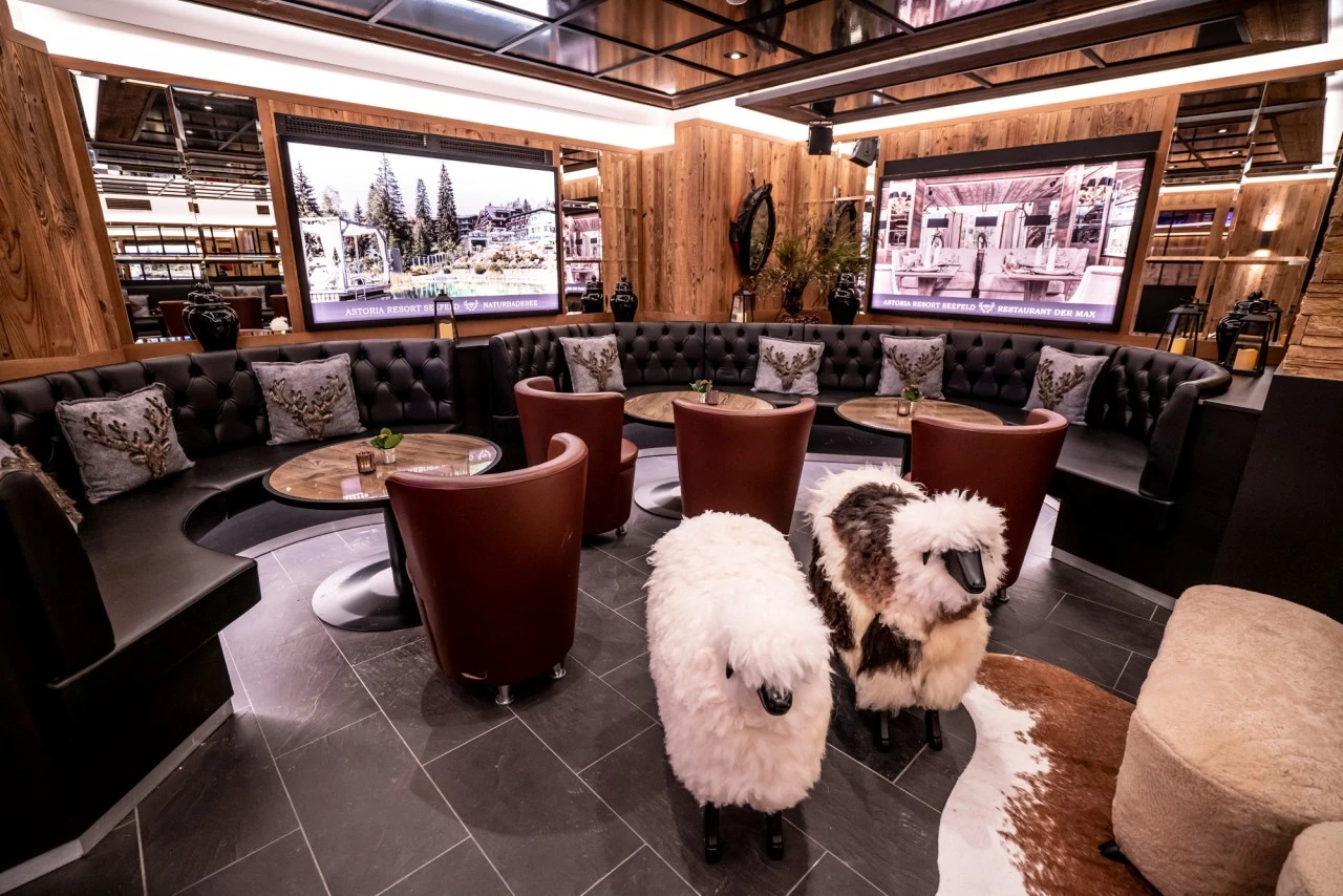 Elegante Lounge mit dunklen Ledersofas, runden Tischen, großen Bildschirmen und zwei dekorativen Schaf-Figuren aus Plüsch.