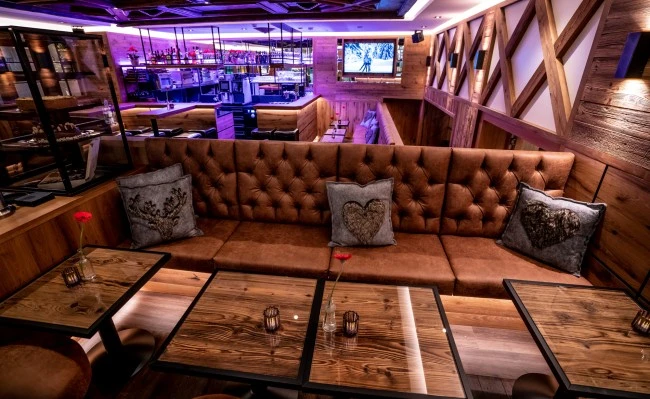 Gemütliche Bar mit rustikalem Holzmobiliar, stimmungsvoller Beleuchtung und dekorativen Kissen mit Herz- und Hirschmotiven.